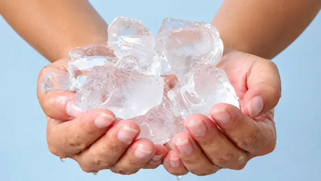 Mãos segurando cubos de gelo usados para tirar cera de depilação do chão antes de raspar com espátula